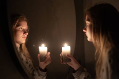 Genç bir kadın aynanın yanında yanan bir mum tutuyor, yansıması ılık parıltıyla hafifçe aydınlatılıyor. Sahne, iç gözlem ve sükunet yayar, barışçıl ve samimi bir atmosfer yaratır.