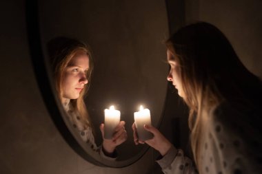 Genç bir kadın yanan bir mum tutarken aynaya bakıyor, sıcak parıltı yüzünü ve yansımasını aydınlatıyor. Huzurlu bir an içgözlem ve sessiz bir zarafeti çağrıştırıyor. Yüksek kalite fotoğraf