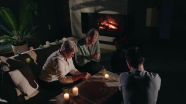 Bir kız, yaşlı annesi mum ışığında bir tarot kartı okurken dikkatlice dinler, sıcak bir oturma odasında sessiz ve mistik bir anı paylaşır. Yüksek kalite 4k görüntü