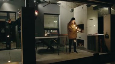 İki adam, akşamları kış tatiline hazırlanmak için bir ev verandasını tel ışıklarıyla süsler. 