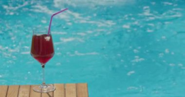 Bir bardak buzlu kırmızı kokteyl ve ahşap bir havuz başında mor bir pipet, güneşli bir yaz gününde arka planda köpüklü mavi su. Yüksek kalite 4k görüntü
