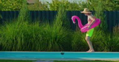 Hasır şapkalı ve neon şortlu genç adam havuz kenarında neşe içinde dans ediyor, pembe flamingo şamandırası tutuyor, eğlenceli bir yaz anının tadını çıkarıyor. Yüksek kalite 4k görüntü