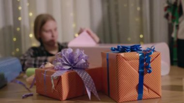 Arka planda ambalaj kağıdı hazırlayan genç bir kız ve mor ve mavi kurdeleli iki güzel paketlenmiş turuncu hediye kutusu. Yüksek kalite 4k görüntü