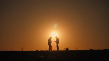 Gün doğumunda ya da günbatımında tarlada iletişim kuran iki siluetli çiftçi. Bir çiftçi elinde tabletle planlarını tartışıyor. Yakınlarda bir kürek ve su kabı görülüyor, takım çalışmasını sembolize ediyor.