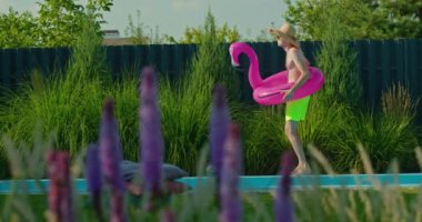Hasır şapkalı ve neon şortlu genç bir adam pembe flamingo şamandıralı bir yüzme havuzunun yanında dans ediyor yemyeşil bir arka bahçede kaygısız bir yaz anının tadını çıkarıyor. Yüksek kalite 4k görüntü