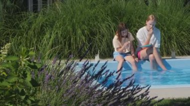 İki genç kız, güneşli bir yaz günü aperatiflerini yerken ve ferahlatıcı içecekler içerken havuz kenarında ayaklarını suya daldırıyorlar. Yüksek kalite 4k görüntü