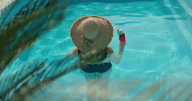 Yüzme havuzunda duran, hasır şapka takan ve elinde tropik bir kokteyl tutan genç bir kadın yaz tatilinin tadını çıkarıyor. Yüksek kalite 4k görüntü
