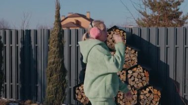 Güneşli bir kış gününde arka bahçesinde dans eden orta yaşlı bir kadın yeşil kapüşonlu ve pembe kulaklıklı. Yığınla odun yığınının önünde neşeyle hareket ediyor. Yüksek kalite 4k görüntü