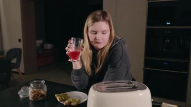 Modern bir mutfakta tost makinesi pişirecek tostu beklerken meyve suyu içen genç bir kadın. Yüksek kalite 4k görüntü