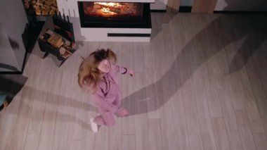 Sıcak bir evde şöminenin yanında dans eden genç bir kadın, yukarıdan izleniyor. Pembe eşofman giyiyor ve beyaz çoraplar giyiyor. Saçları hareket halinde. Ahşap zeminde dramatik bir gölge uzanıyor.