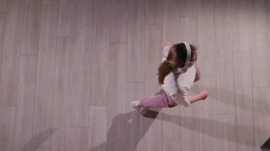 Genç bir kadın, yukarıdan izlenen kulaklıkla müzik dinlerken enerjik bir şekilde dans ediyor. Beyaz bir tişört, pembe eşofman altı ve beyaz çoraplar giyiyor. Hareketi gölgede dinamik bir gölge yaratıyor.