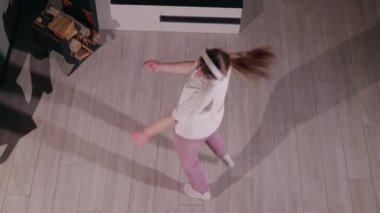 Şöminenin yanında dans eden genç bir kadın, yukarıdan görülüyor. Beyaz bir tişört, pembe eşofman altı ve kulaklıkla ritmik bir şekilde hareket eden beyaz çoraplar giyiyor. Gölgesi ağaçların arasında uzanıyor.