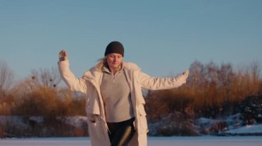 Neşeli orta yaşlı kadın, parlak kış güneşi altında donmuş bir gölde kolları açık dans ediyor. Onun enerjik hareketi özgürlüğü ve mutluluğu yansıtıyor. Yüksek kalite 4k görüntü