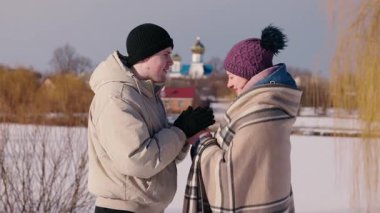 Genç bir adam kış yürüyüşü sırasında kız arkadaşını sıcak bir battaniyeye sarıp karlı ağaçların ve açık mavi gökyüzünün arasında şefkat ve şefkat gösteriyor. Yüksek kalite 4k görüntü