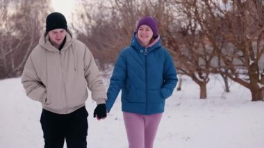 Karlı bir yolda birlikte yürüyen ve gülümseyen genç bir çift hafif kar yağışı sırasında birbirlerine eşlik ederek eğleniyorlar.