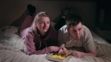 Genç bir çift, modern bir yatakta midelerinin üzerine uzanmış televizyon izliyor ve cips yiyor, fast food atıştırmalıklarıyla rahat bir akşamın tadını çıkarıyor. Yüksek kalite 4k görüntü