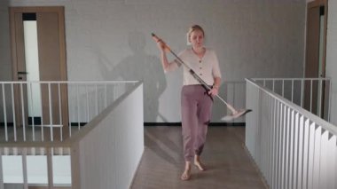 Orta yaşlı bir kadın modern bir koridorda paspasla enerjik bir şekilde dans ediyor, neşe, canlılık ve pozitif enerji saçıyor. Yüksek kalite 4k görüntü