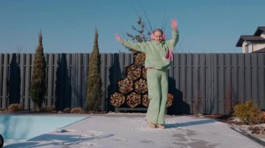 Güneşli bir kış gününde arka bahçesinde kollarını kaldırmış dans eden orta yaşlı bir kadın. Kapalı havuzun yanında yeşil eşofman ve pembe kulaklık giyiyor ve yakacak odunluğu var. Yüksek kalite 4k