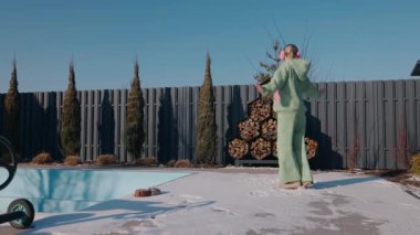Parlak bir kış gününde, karlı bir havuzun yanında dans eden orta yaşlı bir kadın. Yeşil kapüşonlu ve pembe kulaklıklı arka bahçesindeki açık havanın tadını çıkarıyor. Yüksek kalite 4k görüntü