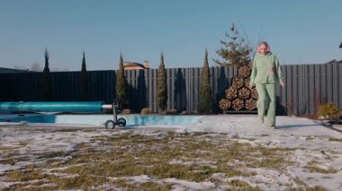 Yeşil eşofmanlı orta yaşlı bir kadın havuz kenarındaki karlı bir terasta neşeyle zıplıyor, temiz kış havasının tadını çıkarıyor. Yüksek kalite 4k görüntü