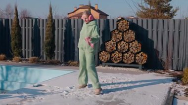 Güneşli bir kış gününde arka bahçesinde dans eden orta yaşlı bir kadın yeşil eşofman ve pembe kulaklık giyiyor. Kapalı bir havuzun yanındaki karlı zeminde neşeyle hareket ediyor ve yakacak odun yığıyor. Yüksek