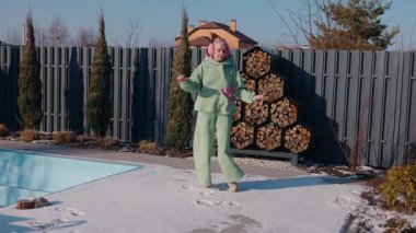 Güneşli bir kış gününde arka bahçesinde dans eden orta yaşlı bir kadın. Kulaklıklı, sıcak yeşil bir kıyafet giyiyor. Kar, üstü örtülü bir havuz ve yakacak odun rafının yakınındaki zemini kaplıyor. Yüksek kalite 4k
