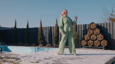 Yeşil eşofmanlı orta yaşlı bir kadın havuz kenarında karlı bir verandada dans ediyor, açık havada açık kış gününün tadını çıkarıyor. Yüksek kalite 4k görüntü
