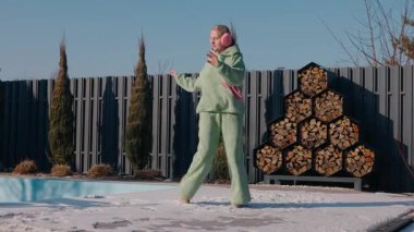 Parlak bir kış gününde arka bahçesinde enerjik bir şekilde dans eden orta yaşlı bir kadın. Kapalı havuzun yanında pembe kulaklıklı yeşil bir kapüşonlu ve düzgünce dizilmiş odun askısı var. Yüksek kalite 4k