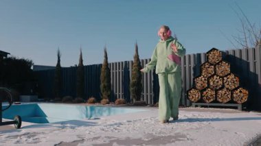 Güneşli bir kış gününde karlı arka bahçesinde dans eden orta yaşlı bir kadın. Kapalı havuzun yanında yeşil bir kapüşonlu ve pembe bir kulaklık takıyor ve odun yığıyor. Açık havanın tadını çıkarıyor. Yüksek kalite 4k