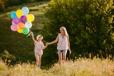 Orta yaşlı bir kadın renkli balonlar taşıyan neşeli bir kızla el ele tutuşuyor yemyeşil yemyeşil bir çayırda yürüyor. Yüksek kalite fotoğraf