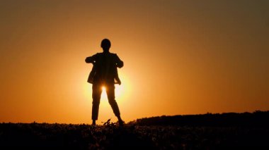 Gün batımında kırsal bir alanda zarif bir şekilde dans eden orta yaşlı bir kadın silueti, dingin ve anlamlı bir hareketle altın gökyüzü ile harmanlanıyor. Yüksek kalite 4k görüntü