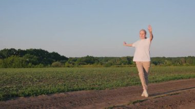Kırsal bir alanda, toprak bir yolda coşkuyla dans eden orta yaşlı bir kadın, doğadaki neşeli ve tasasız bir anı kucaklıyor. Yüksek kalite 4k görüntü