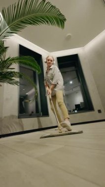 Orta yaşlı bir kadın, yeri paspaslarken ve kulaklıkla müzik dinlerken modern bir odada, saksıda avuç içiyle eğleniyor. Yüksek kalite 4k görüntü