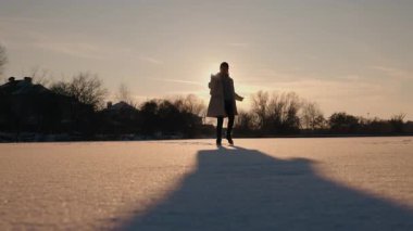 Orta yaşlı bir kadın gün batımında donmuş bir gölde zarif bir şekilde dans ediyor, güneş arkadan vuruyor. Uzun gölgeler ve altın ışık büyülü ve huzurlu bir atmosfer yaratır. Yüksek kalite 4k görüntü