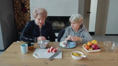 İki yaşlı kadın, sıcak bir ev mutfağında, ahşap bir masada birlikte yemek hazırlıyorlar. Yüksek kalite 4k görüntü