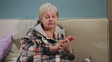 Yatakta oturan yaşlı bir kadın, sıcak ekose bir kapüşon giyerken cildine el kremi sürerek huzurlu bir akşam rutininin tadını çıkarıyor. Yüksek kalite 4k görüntü