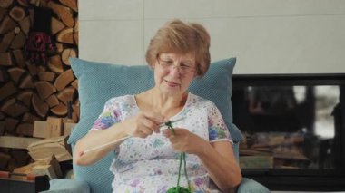 Yesil iplikle örgü ören yasli bir kadin şöminenin yanindaki rahat bir koltukta yakacak odunlu oturuyor. Yüksek kalite 4k görüntü