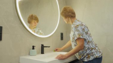 Modern bir banyo lavabosunu süngerle temizleyen yaşlı bir kadın, yansıması yuvarlak bir LED aynada görülebiliyor. Yüksek kalite 4k görüntü