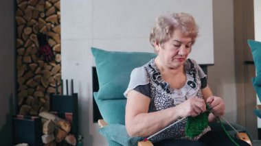 Yeşil iplikle örgü ören yaşlı bir kadın şöminenin yanındaki rahat koltukta otururken işine odaklanmış. Yüksek kalite 4k görüntü