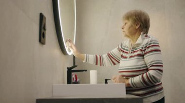 Modern bir banyoda yuvarlak ışıklı aynayı temizleyen, çizgili bir kazak giyen yaşlı bir kadın. Yüksek kalite 4k görüntü