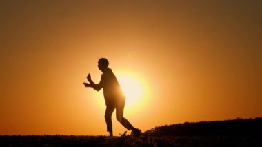 Gün batımında kırsal bir alanda hayali davul çalan orta yaşlı bir kadın, silueti anlamlı bir performansla parlayan akşam gökyüzüne karşı duruyor. Yüksek kalite 4k görüntü