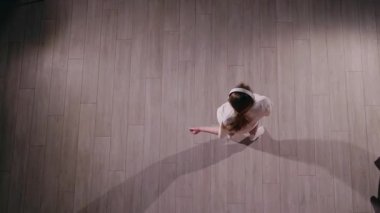 Enerjik genç bir kadın kulaklıkla müzik eşliğinde dans ediyor, yukarıdan çekiliyor. Beyaz bir tişört, pembe eşofman altı ve beyaz çoraplar giyer, dinamik bir gölge oluştururken zarafetle döner. Yüksek
