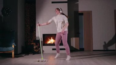 Temizlik yaparken, kulaklık takmış, neşeyle dans eden genç bir kadın. Beyaz tişört, pembe eşofman ve beyaz çorap giyer ev işlerini eğlenceli ve enerjik bir ana çevirir.