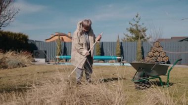 Bahçedeki kuru yaprakları tırmıklayan yaşlı bir kadın bahar hazırlığı için çimleri temizliyor. Yüksek kalite 4k görüntü