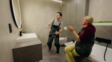 Oyuncu teknisyen banyoda enerjik bir şekilde dans ederken, bir kadın neşeyle alkışlıyor, eğlenceli ve neşeli bir anı paylaşıyor. Yüksek kalite 4k görüntü