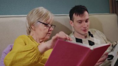 Yaşlı kadın konsantre bir şekilde kitap okurken yanında genç bir adam gülümsüyor, evde rahat bir anın tadını çıkarıyor. Yüksek kalite 4k görüntü