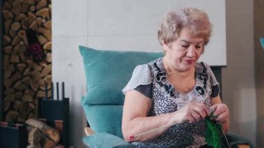 Yeşil iplikle örgü ören yaşlı bir kadın şöminenin yanındaki rahat koltukta huzurlu bir ifadeyle sanatına konsantre oluyor. Yüksek kalite 4k görüntü