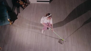 Temizlik yaparken paspasla dans eden genç bir kadın, yukarıdan izlenmiş. Beyaz bir tişört, pembe eşofman altı ve beyaz çoraplar giyiyor, kulaklıkla müzik dinliyor ve ev işlerini eğlenceli hale getiriyor.