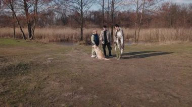 Yaşlı bir kadın, iki yetişkin torunu ve serin bir bahar gününde manzaralı bir gölün yanında duran bir Golden Retriever. Sıcak giyinmişler, ağaçlar ve sazlıklar eşliğinde huzurlu doğal ortamın tadını çıkarıyorlar.