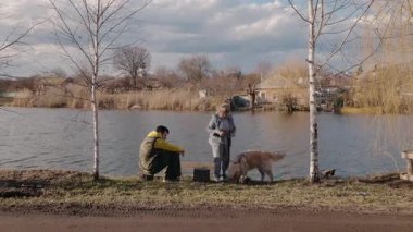 Yaşlı bir kadın, Golden Retriever 'ını nehir kenarında gezdirirken torunu da bahar güneşinin altında bir ağaç kütüğünde dinleniyor. Arka planda köy ve kilise görünüyor.